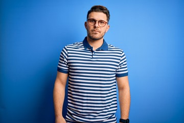 Young man with blue eyes wearing glasses and casual striped t-shirt over blue background Relaxed with serious expression on face. Simple and natural looking at the camera.