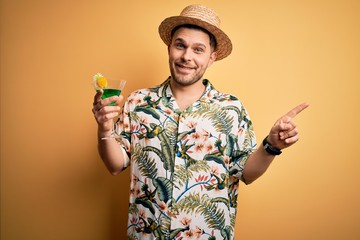 Young man on vacation wearing summer hat drinking a party cocktail over yellow background very happy pointing with hand and finger to the side