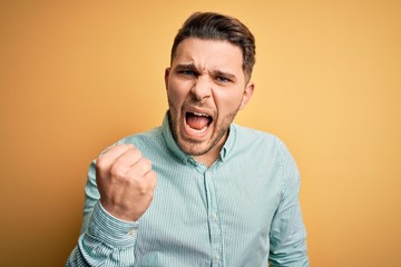 Obraz premium Young business man with blue eyes wearing elegant green shirt over yellow background angry and mad raising fist frustrated and furious while shouting with anger. Rage and aggressive concept.