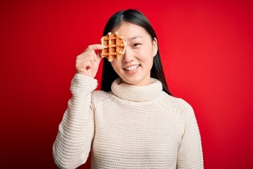 Young asian woman eating sweet and tasty belgian waffle over red isolated background with a happy face standing and smiling with a confident smile showing teeth