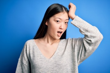 Fototapeta premium Young beautiful asian woman wearing casual sweater standing over blue isolated background surprised with hand on head for mistake, remember error. Forgot, bad memory concept.