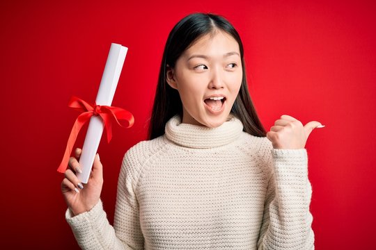 Young asian student woman holding graduate diploma over red isolated background pointing and showing with thumb up to the side with happy face smiling