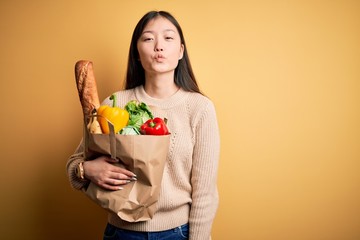 Young asian woman holding paper bag of fresh healthy groceries over yellow isolated background looking at the camera blowing a kiss on air being lovely and sexy. Love expression.