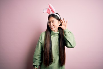 Young asian woman wearing cute easter bunny ears over pink background smiling with hand over ear listening an hearing to rumor or gossip. Deafness concept.