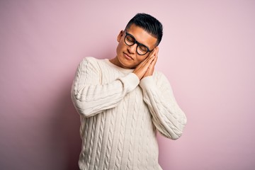 Fototapeta premium Young handsome latin man wearing white casual sweater and glasses over pink background sleeping tired dreaming and posing with hands together while smiling with closed eyes.