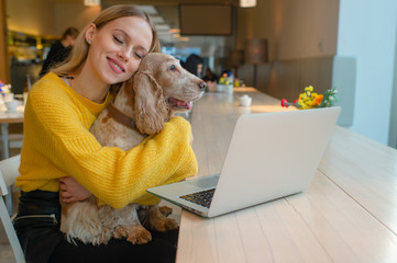 Happy smiling blonde woman hugging her lovely cocker spaniel puppy with eyes closed and joyful face expression in front of laptop on the table in caffe.