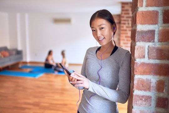 Young beautiful chinese sportswoman smiling happy. Standing listening to music using earphones and smartphone after class of yoga at gym