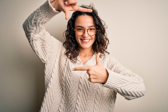 Beautiful woman with curly hair wearing casual sweater and glasses over white background smiling making frame with hands and fingers with happy face. Creativity and photography concept.