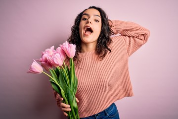Young beautiful romantic woman with curly hair holding bouquet of pink tulips Crazy and scared with hands on head, afraid and surprised of shock with open mouth