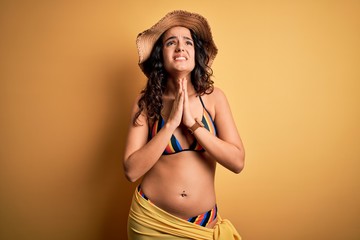 Young beautiful woman with curly hair on vacation wearing bikini and summer hat begging and praying...