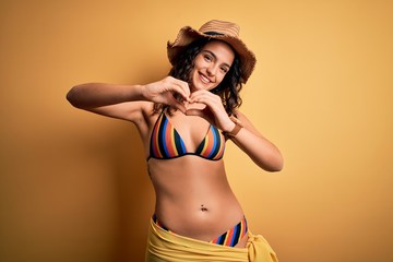 Young beautiful woman with curly hair on vacation wearing bikini and summer hat smiling in love doing heart symbol shape with hands. Romantic concept.