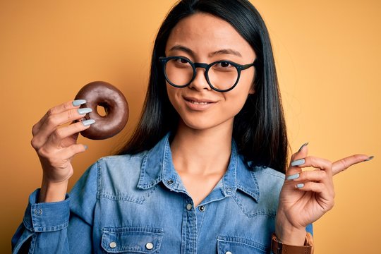 Young beautiful chinese woman holding chocolate donut over isolated yellow background very happy pointing with hand and finger to the side