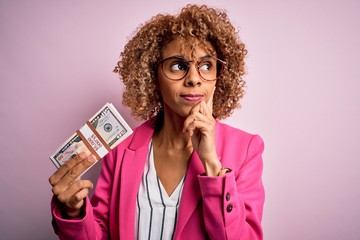 Young african american curly woman holding bunch of dollars banknotes over pink background serious...