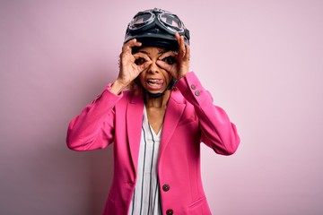 African american motorcyclist woman with curly hair wearing moto helmet over pink background doing ok gesture like binoculars sticking tongue out, eyes looking through fingers. Crazy expression.