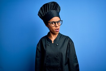 Young african american chef woman wearing cooker uniform and hat over blue background In shock face, looking skeptical and sarcastic, surprised with open mouth