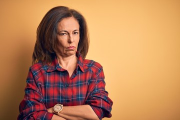 Middle age beautiful woman wearing casual shirt standing over isolated yellow background skeptic and nervous, disapproving expression on face with crossed arms. Negative person. - obrazy, fototapety, plakaty