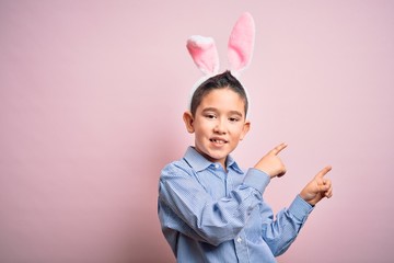Young little boy kid wearing easter bunny ears over isolated pink background smiling and looking at the camera pointing with two hands and fingers to the side.