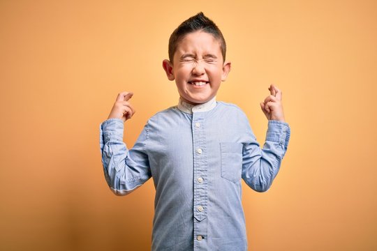Young little boy kid wearing elegant shirt standing over yellow isolated background gesturing finger crossed smiling with hope and eyes closed. Luck and superstitious concept.