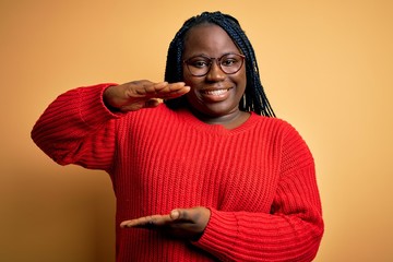 African american plus size woman with braids wearing casual sweater over yellow background gesturing with hands showing big and large size sign, measure symbol. Smiling looking at the camera. Measure