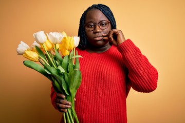 Young african american plus size woman with braids holding bouquet of yellow tulips flower mouth...