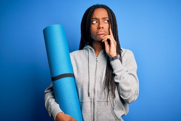 Young african american fitness woman holding yoga mat over isolated blue background serious face...