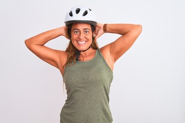 Middle age mature cyclist woman wearing safety helmet over isolated background relaxing and stretching, arms and hands behind head and neck smiling happy