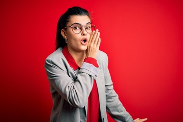 Young beautiful brunette businesswoman wearing jacket and glasses over red background hand on mouth telling secret rumor, whispering malicious talk conversation