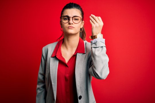 Young beautiful brunette businesswoman wearing jacket and glasses over red background Doing Italian gesture with hand and fingers confident expression