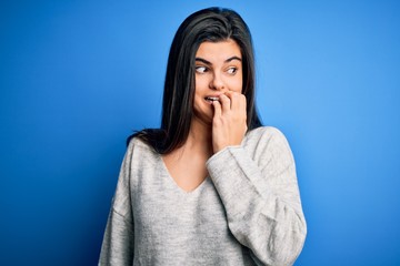 Young beautiful brunette woman wearing casual sweater standing over blue background looking stressed and nervous with hands on mouth biting nails. Anxiety problem.