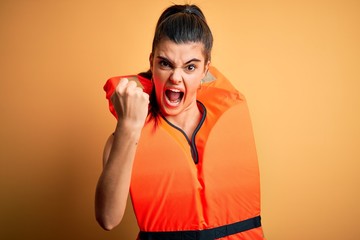 Young beautiful brunette woman wearing orange safe lifejacket over yellow background angry and mad...