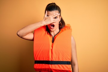 Young beautiful brunette woman wearing orange safe lifejacket over yellow background peeking in...