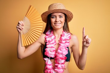 Young beautiful woman with blue eyes on vacation wearing bikini holding hand fan surprised with an idea or question pointing finger with happy face, number one