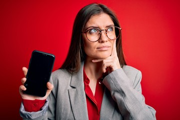 Young beautiful business woman with blue eyes holding smartphone showing screen serious face...