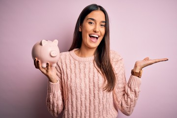 Young beautiful brunette woman holding piggy bank saving money for retirement very happy and...