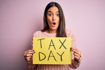 Young beautiful brunette woman holding paper with tax day message over pink background scared in...