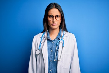 Young beautiful doctor woman wearing stethoscope and glasses over blue background skeptic and nervous, frowning upset because of problem. Negative person.