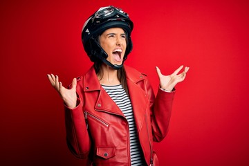 Young beautiful brunette motorcyclist woman wearing motorcycle helmet and red jacket crazy and mad shouting and yelling with aggressive expression and arms raised. Frustration concept.