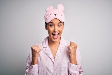 Young beautiful brunette woman wearing pajama and sleep mask over white background celebrating surprised and amazed for success with arms raised and open eyes. Winner concept.