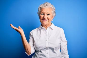 Senior beautiful woman wearing elegant shirt standing over isolated blue background smiling cheerful presenting and pointing with palm of hand looking at the camera.