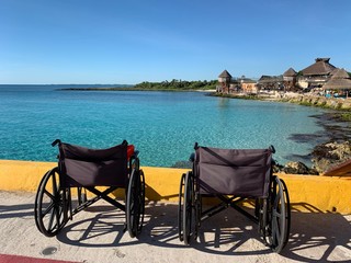 Two wheelchairs are on the pier in Costa Maya, Mexico. Accessible for the cruise ship guests.  Blue sea is on a background. View on a thatched roofs and coral reef.