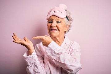 Senior beautiful woman wearing sleep mask and pajama over isolated pink background amazed and smiling to the camera while presenting with hand and pointing with finger.