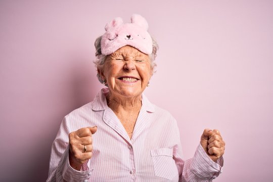 Senior beautiful woman wearing sleep mask and pajama over isolated pink background excited for success with arms raised and eyes closed celebrating victory smiling. Winner concept.