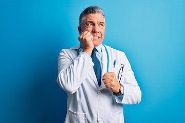 Middle age handsome grey-haired doctor man wearing coat and blue stethoscope looking stressed and nervous with hands on mouth biting nails. Anxiety problem.
