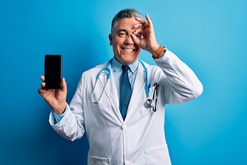 Middle age handsome grey-haired doctor man holding smartphone showing screen with happy face smiling doing ok sign with hand on eye looking through fingers