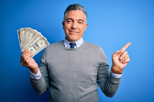 Middle age handsome grey-haired man holding bunch of dollars over blue background very happy pointing with hand and finger to the side