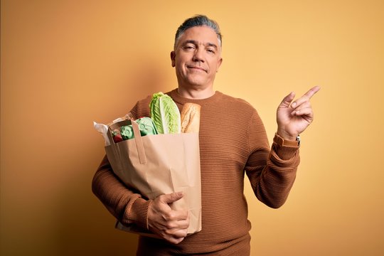 Middle age handsome grey-haired man holding paper bag with food over yellow background very happy pointing with hand and finger to the side