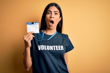 Young beautiful hispanic woman wearing volunteer t-shirt and showing identification scared in shock with a surprise face, afraid and excited with fear expression