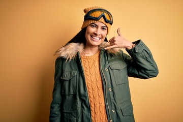 Young beautiful hispanic woman wearing ski glasses and coat for winter weather smiling doing phone gesture with hand and fingers like talking on the telephone. Communicating concepts.