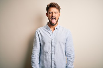 Young handsome man with beard wearing striped shirt standing over white background sticking tongue out happy with funny expression. Emotion concept.