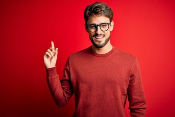Naklejka premium Young handsome man with beard wearing glasses and sweater standing over red background with a big smile on face, pointing with hand finger to the side looking at the camera.